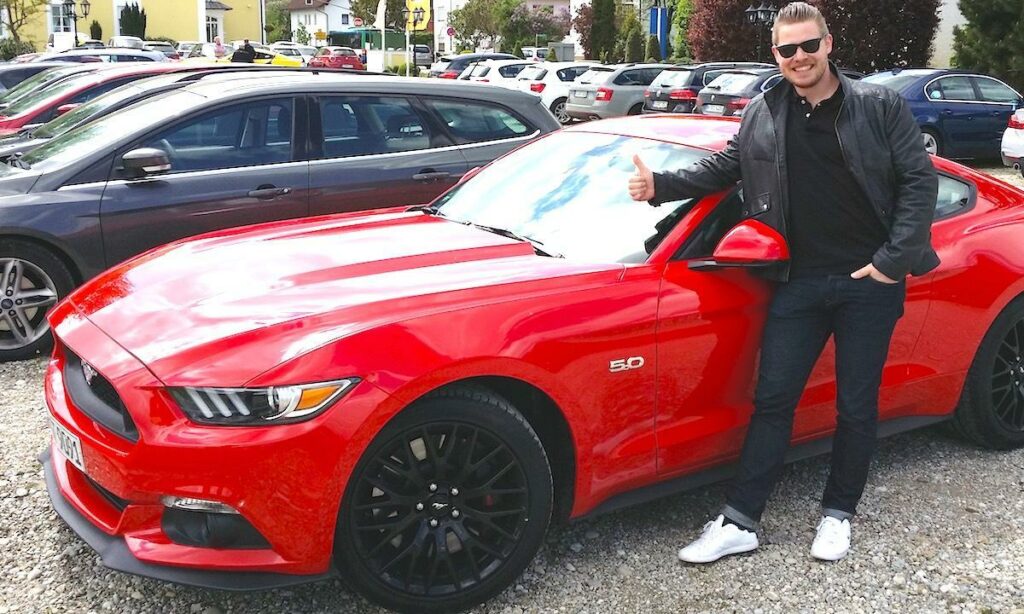 En man poserar bredvid en röd Ford Mustang. (AI text)
