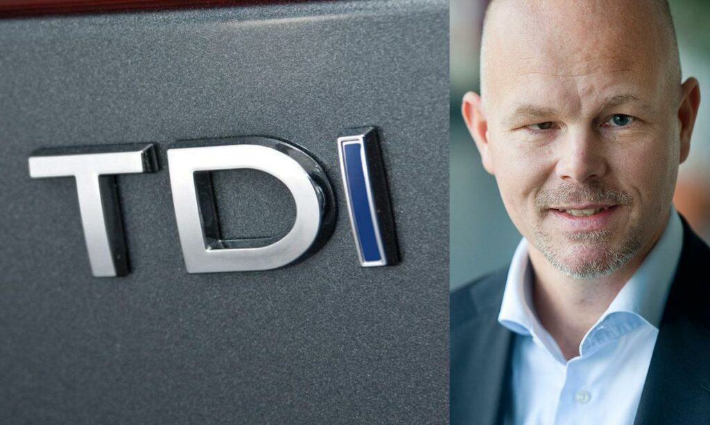 En man i kostym och slips står bredvid ett TDI-logotyp. (AI text)