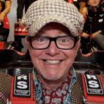 En man i hatt och glasögon poserar tillsammans med en racebil. (AI text)