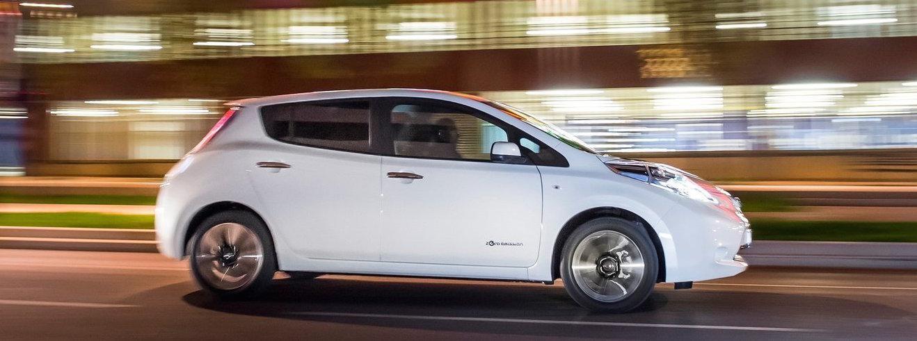 En vit Nissan Leaf kör nedför en stadsgata på natten. (AI text)
