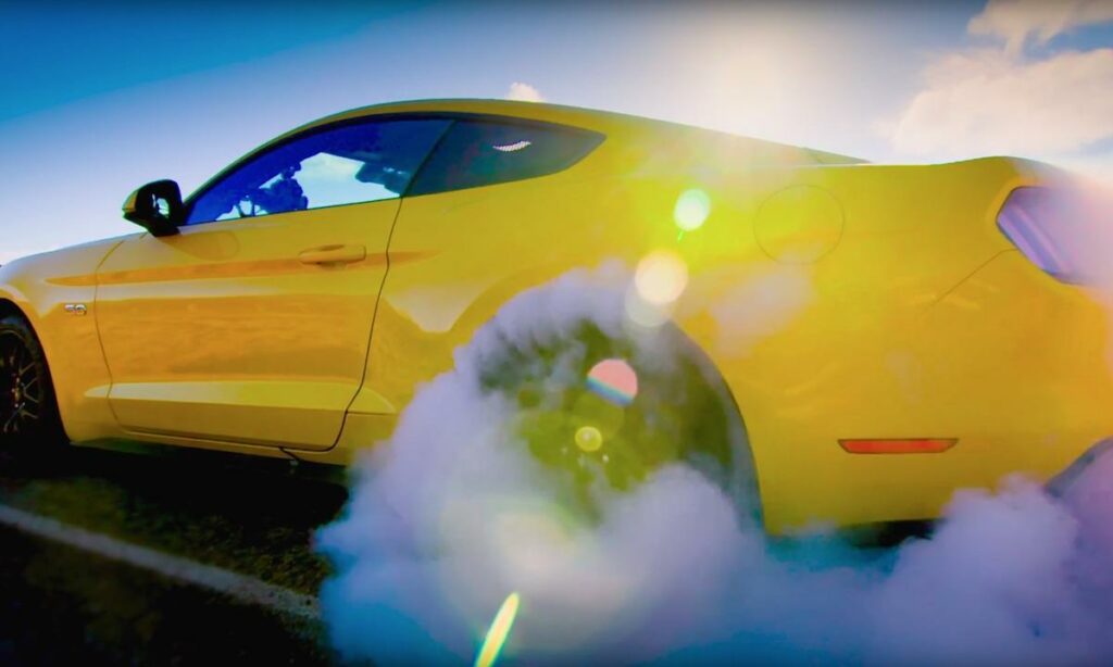 En gul Mustang kör nedför vägen med rök som kommer utifrån den. (AI text)