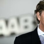 En man i kostym och slips talar till en kamera. (AI text)