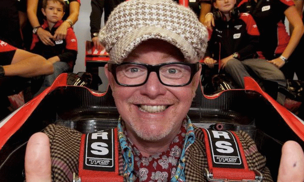 En man i hatt och glasögon poserar tillsammans med en racebil. (AI text)