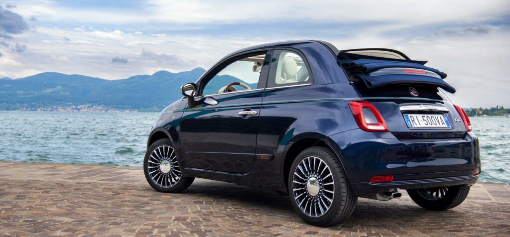 En Fiat 500 cabriolet parkerad på en gata mittemot en sjö. (AI text)