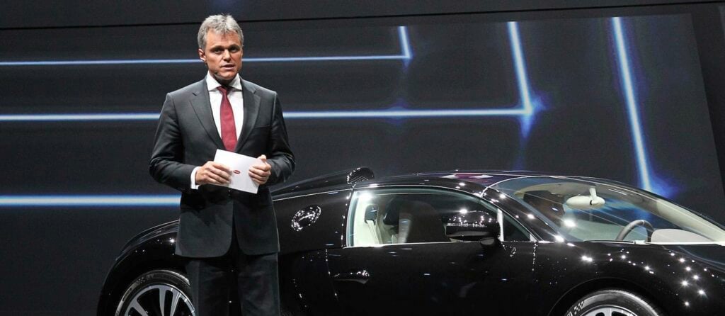 En man i kostym står bredvid en svart Bugatti Veyron på bilsalongen. (AI text)