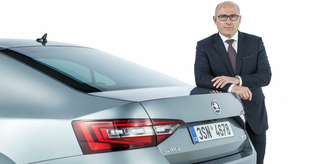 En man i kostym står bredvid en silverfärgad bil. (AI text)