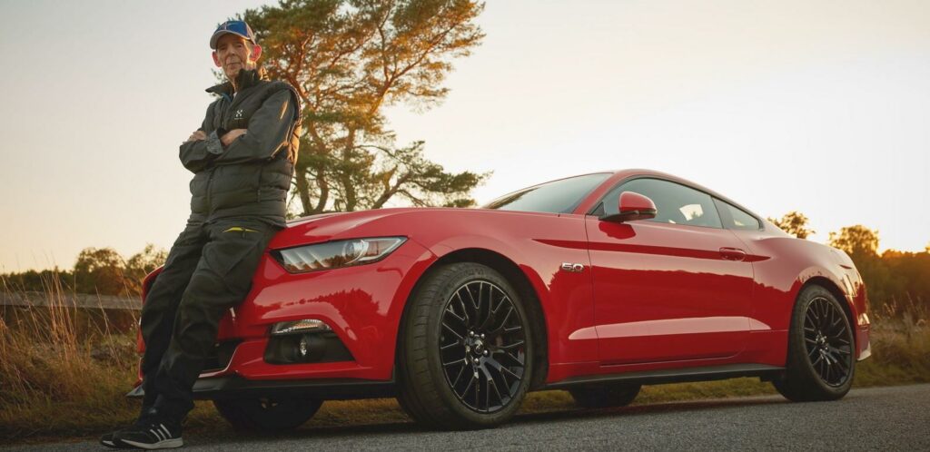 En man står bredvid en röd Ford Mustang. (AI text)