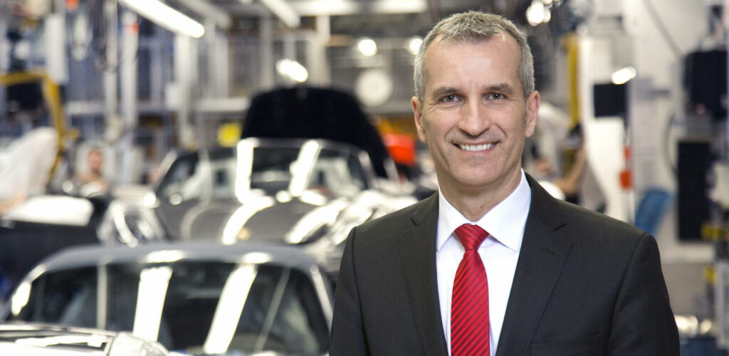 En man i kostym står framför en biltillverkning. (AI text)