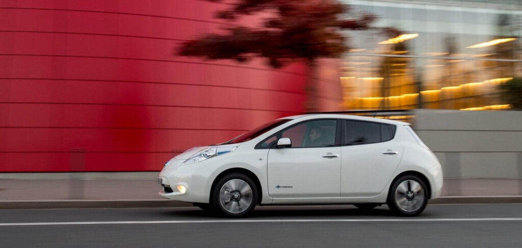 En vit Nissan Leaf kör nedför en gata. (AI text)