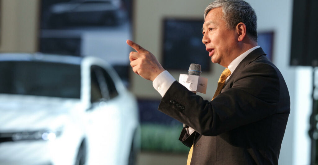 En man i kostym håller en tal vid en bilmesse. (AI text)