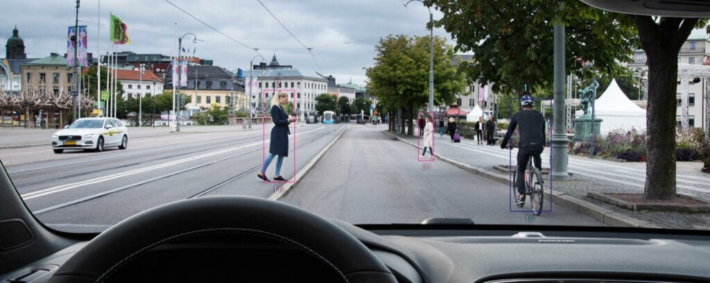 En bil kör framför en gata med människor på trottoaren. (AI text)