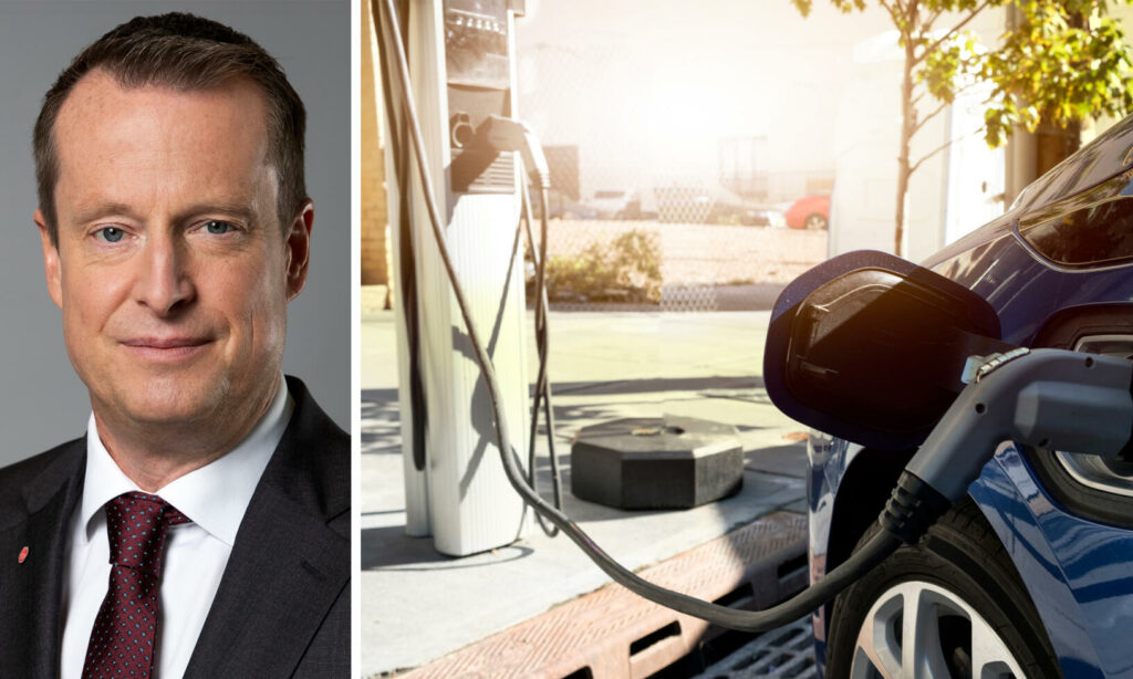 En man i kostym och slips står bredvid en bil som laddas på en elbilstation. (AI text)