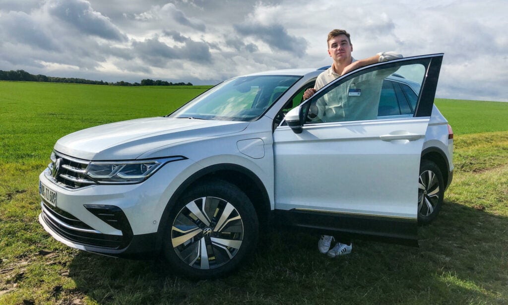 En person står bredvid en vit Volkswagen Tiguan i en åker. (AI text)