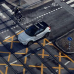 Övervakningsvy av en stadsgata med bilar och fotgängare som korsar gatan. (AI text)