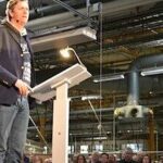 En man talar till en skara människor på en fabrik. (AI text)