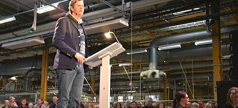 En man talar till en skara människor på en fabrik. (AI text)