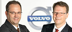 Två män i kostym står bredvid en Volvo-logotyp. (AI text)