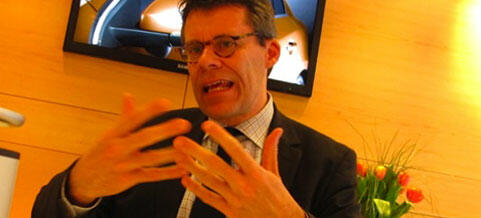 En man i kostym och slips pratar inför en kamera. (AI text)