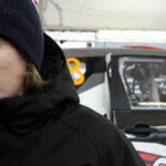 En man i en mössa står bredvid en rallybil. (AI text)