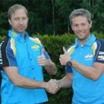 Två män i blå skjortor står bredvid en bil. (AI text)