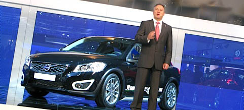 En man i kostym står på en scen bredvid en bil. (AI text)