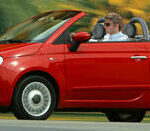 En röd Fiat 124 cabriolet kör nedför en väg. (AI text)