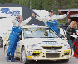 En rallybil med två personer stående på den. (AI text)