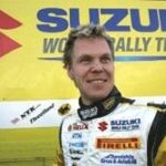 En man i gul klädsel står framför en Suzuki WRC-teammärke. (AI text)