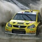 En gul Suzuki Rally-bil kör genom en smutsig yta. (AI text)
