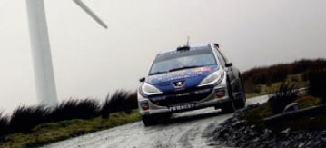 En rallybil körer nedför en väg med vindkraftverk i bakgrunden. (AI text)