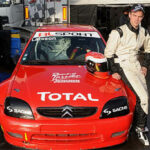 En man i racerutstyrsel står bredvid en röd bil. (AI text)