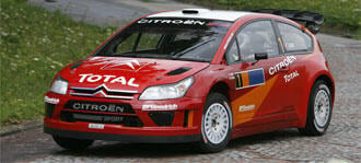 En röd Citroën C3 rallybil kör nedför en väg. (AI text)