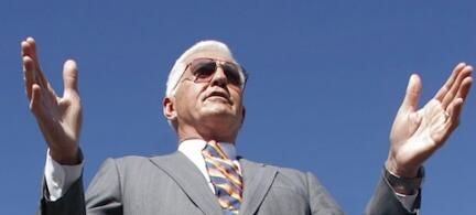 En man i kostym och slips vinkar med händerna i luften. (AI text)