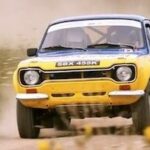 En gul och vit rallybil kör nerför en grusväg. (AI text)