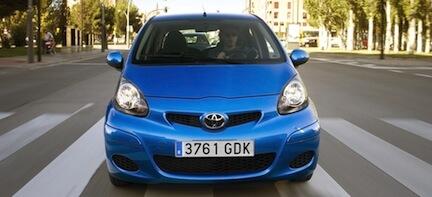 En blå Toyota Aygo kör nedför en gata. (AI text)