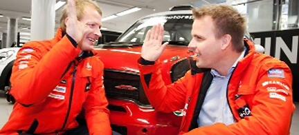 Två män i röda jackor står bredvid en röd Mini. (AI text)