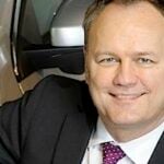 En man i kostym lutar sig mot en bil. (AI text)