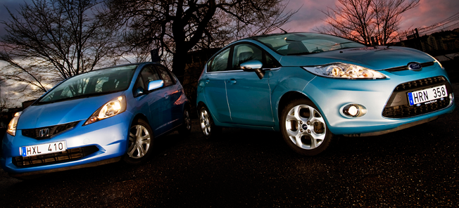 En blå Honda Fusion och en blå Honda Fit parkerade på en gata. (AI text)