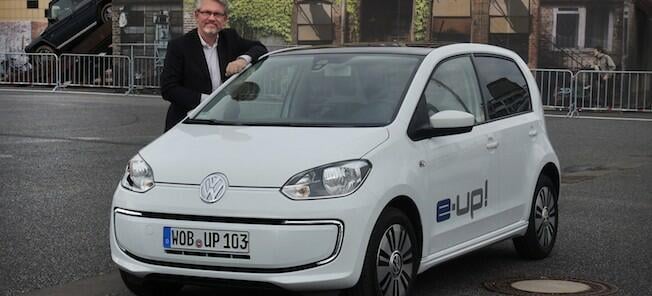 En man står bredvid en vit Volkswagen up! (AI text)