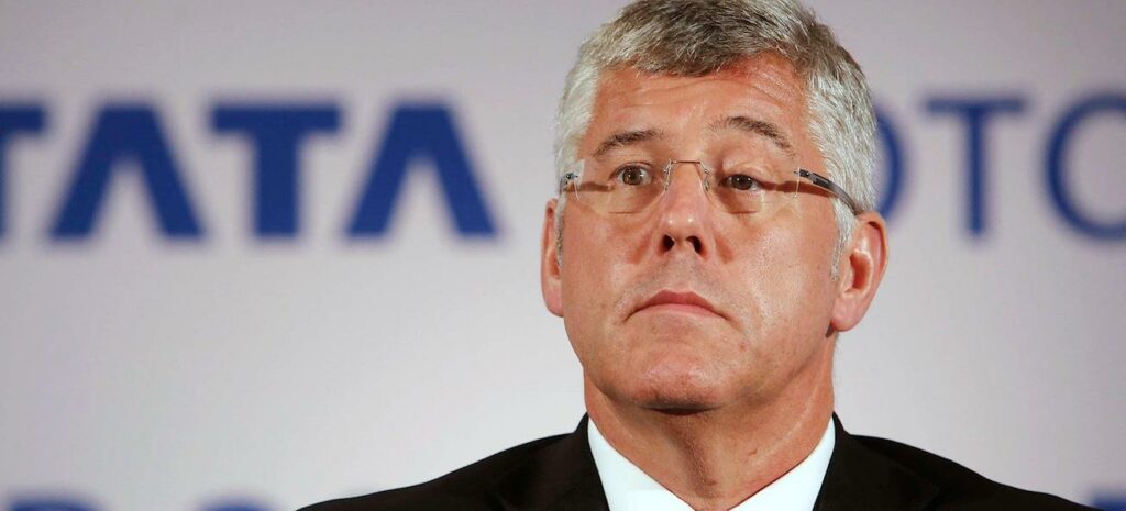 En man i kostym och slips sitter framför TATA-logotypen. (AI text)