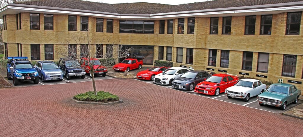 En parkeringsplats med bilar parkerade framför ett byggnad. (AI text)