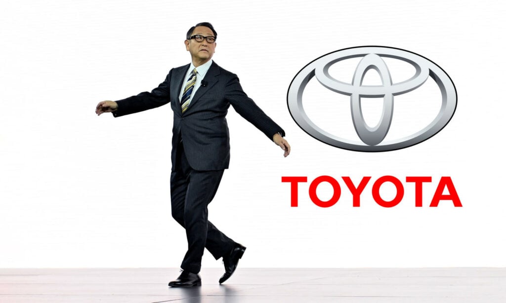 En man i kostym och slips dansar framför Toyotas logotyp. (AI text)