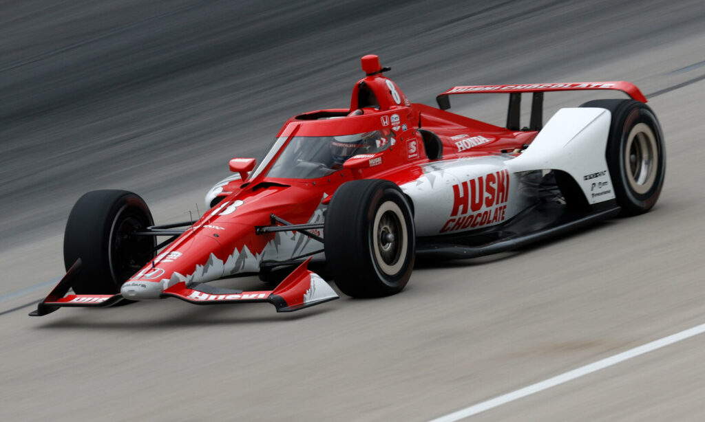 En röd och vit Indy-car tävlar ner på en bana. (AI text)