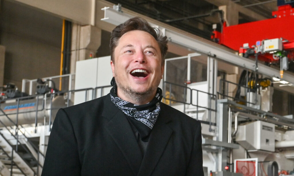 Elon Musk le är en glimt i ansiktet bredvid en fabrik. (AI text)