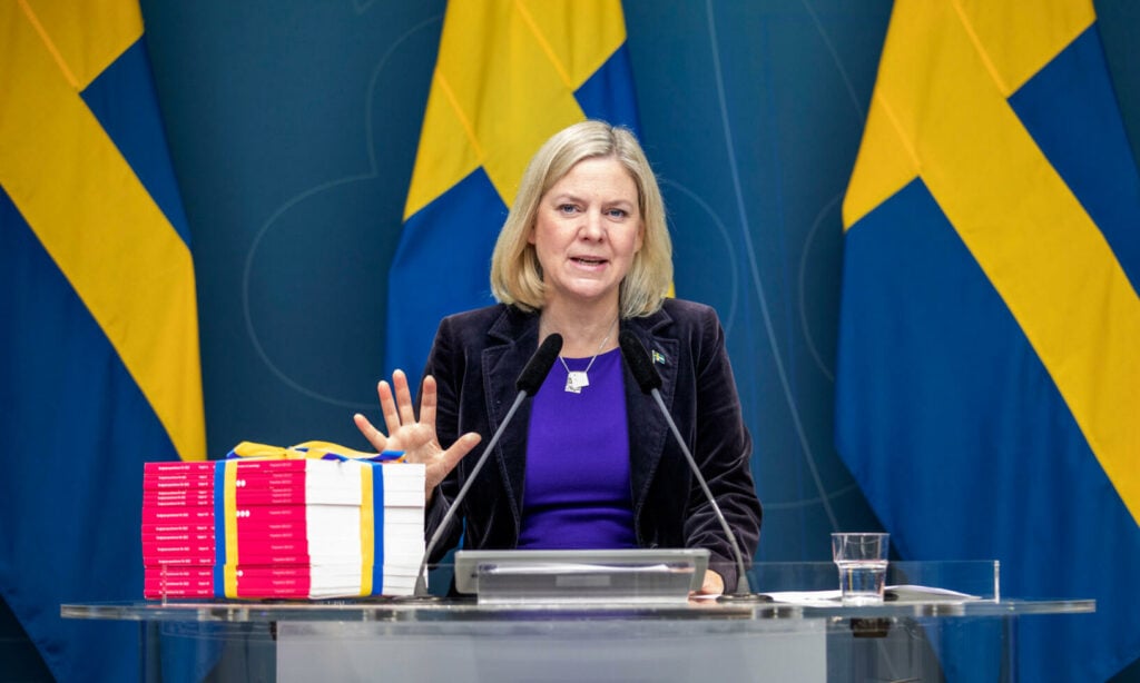 En kvinna står vid ett podiet framför svenska flaggor. (AI text)