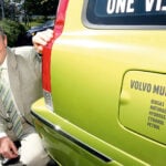 En man i kostym står och poserar bredvid en grön bil. (AI text)