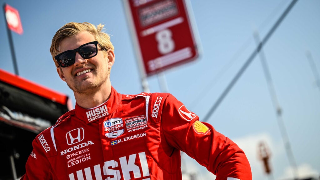 En racerförare i en röd uniform står bredvid en bil. (AI text)