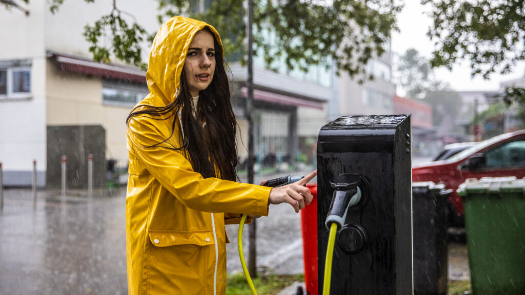 En kvinna i en gul gummiställ klär sin elbil på en laddstation under regn. (AI text)