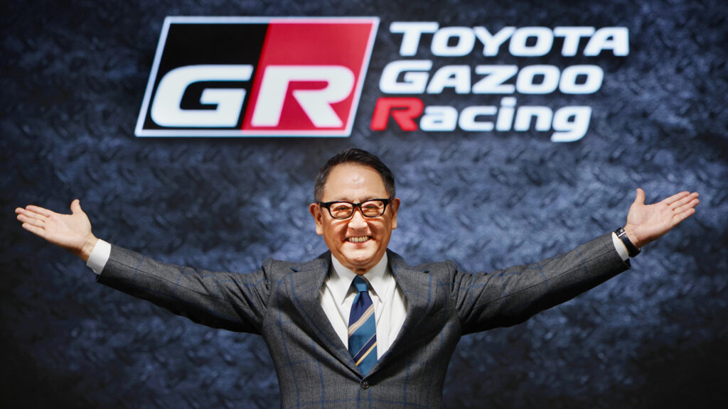 En man i kostym står framför Toyotas GAZOO Racing-logotyp. (AI text)