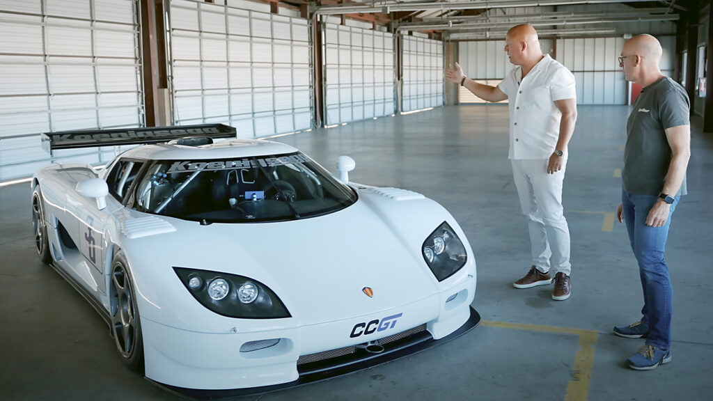 En vit sportbil i en garagem med två män stående bredvid den. (AI text)